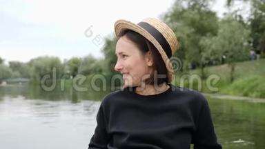 快乐害羞的女孩戴着草帽在城市公园的湖边休息。 年轻的黑发女孩害羞和微笑。 戴着复古帽子的漂亮女孩